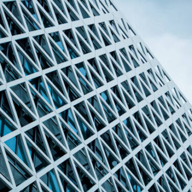 This captivating image showcases a modern business building adorned with a sleek glass facade with metal triangles, standing proudly in the enchanting daylight blue light. The gentle, cool hues of the sky cast a soft and serene glow upon the structure, creating an atmosphere of tranquility and sophistication. The building's reflective glass exterior mirrors the surrounding sky environment, creating a mesmerizing interplay of light and reflections. This image serves as a stunning abstract background, exuding a sense of contemporary elegance and professional allure, making it ideal for a wide range of design and business applications. Copy space.